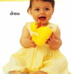 A baby in a yellow dress holding a large yellow rubber duck, sitting on the floor and smiling.