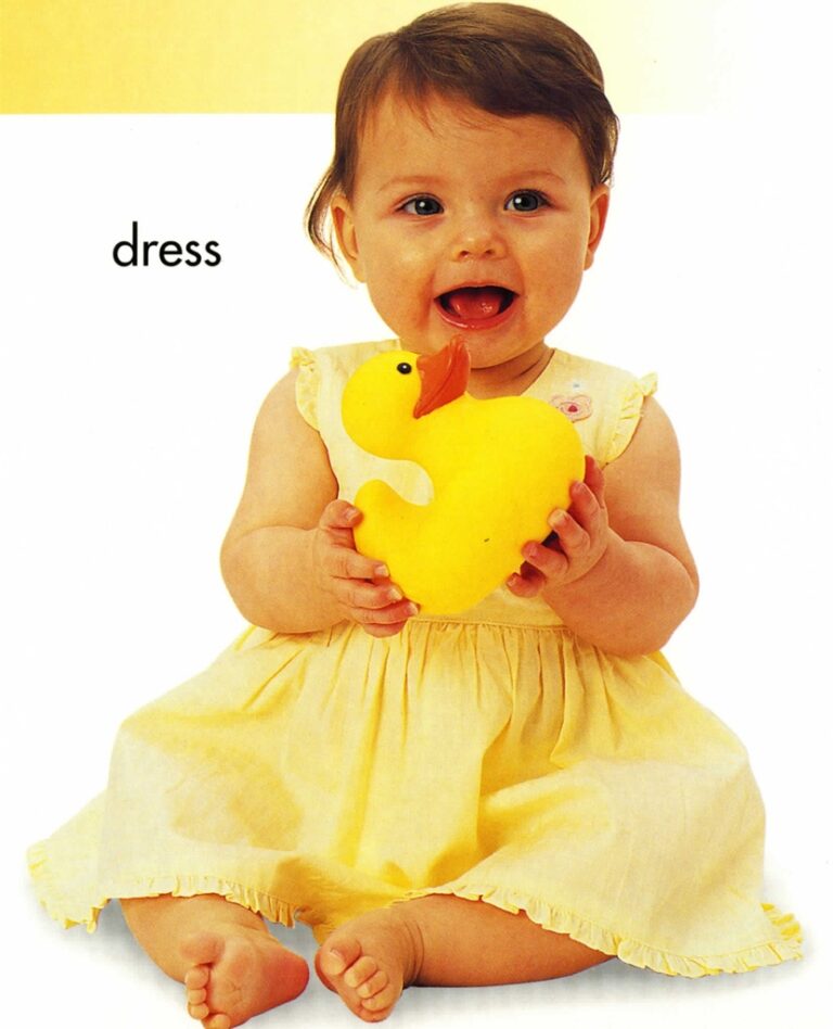 A baby in a yellow dress holding a large yellow rubber duck, sitting on the floor and smiling.